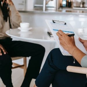 Two people are sitting down. One person is holding and writing on a clipboard and the other person remains sitting in a thoughtful position, with one hand to their face and the other positioned on their thigh.